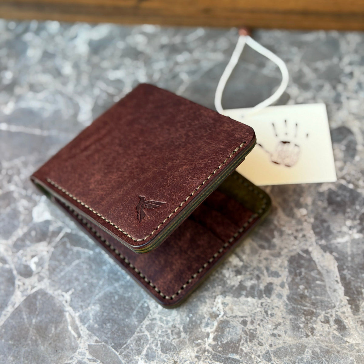A dark brown leather wallet with a matching paper tag attached, resting on a gray marble surface with a wooden background.