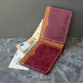 A personalized leather wallet in two-tone red and brown, shown open with a 50 Euro note tucked inside, set against a gray marble surface and textured gray background.