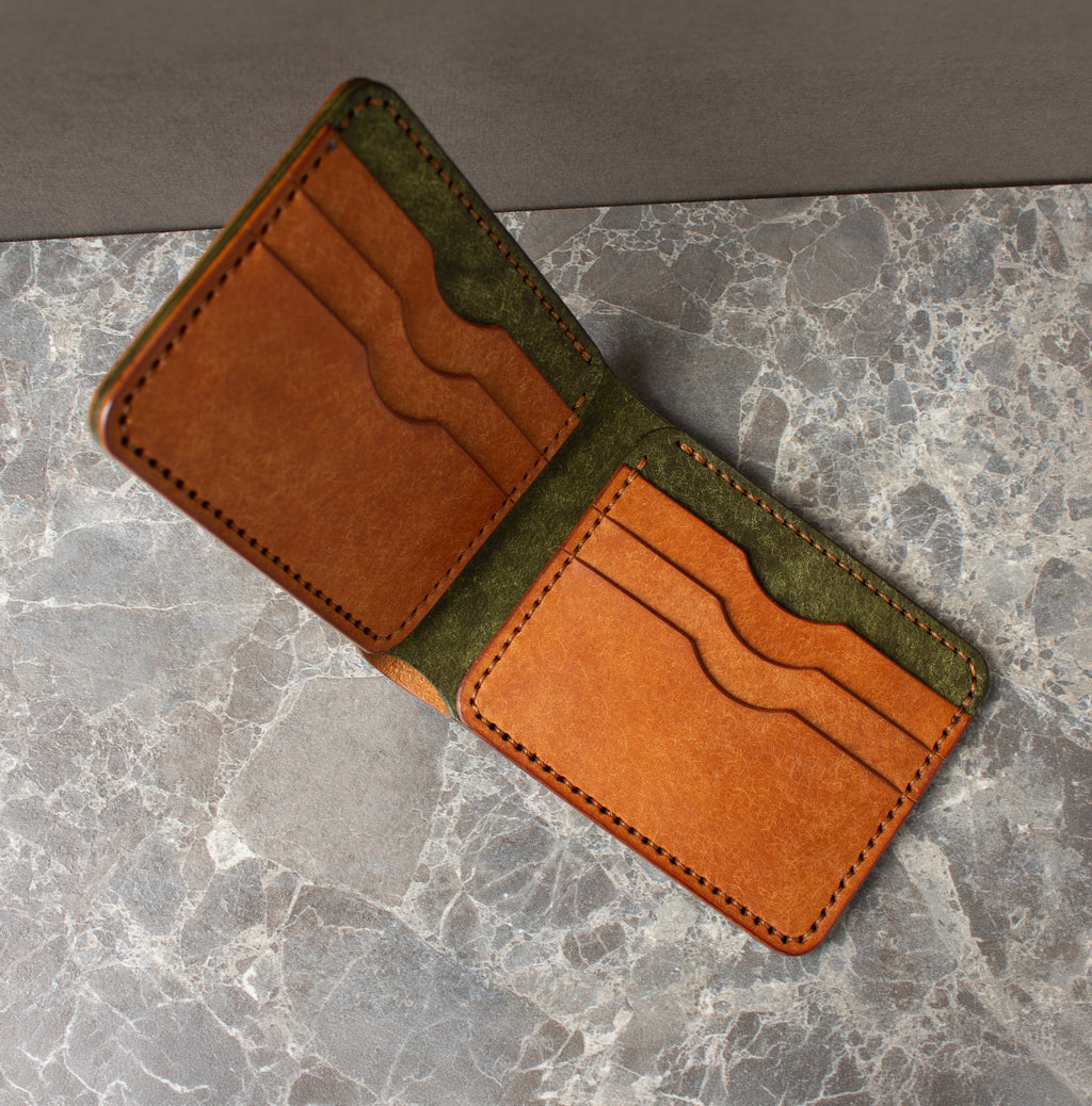 An open bifold wallet made of tan leather with an olive green suede-like lining, displayed diagonally on a gray marble surface.