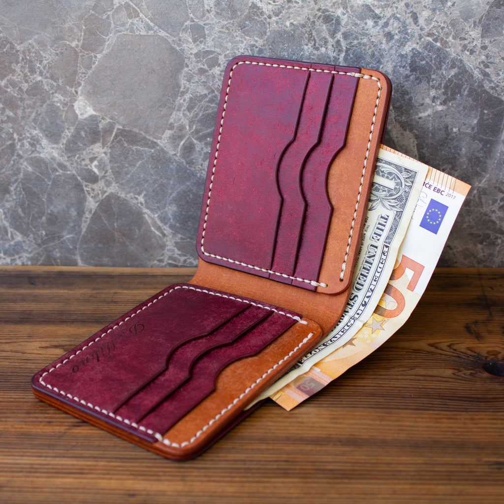 A two-tone leather card holder wallet in burgundy and tan, shown open with Euro banknotes tucked into the card slots, set on a wooden surface against a gray marble background.