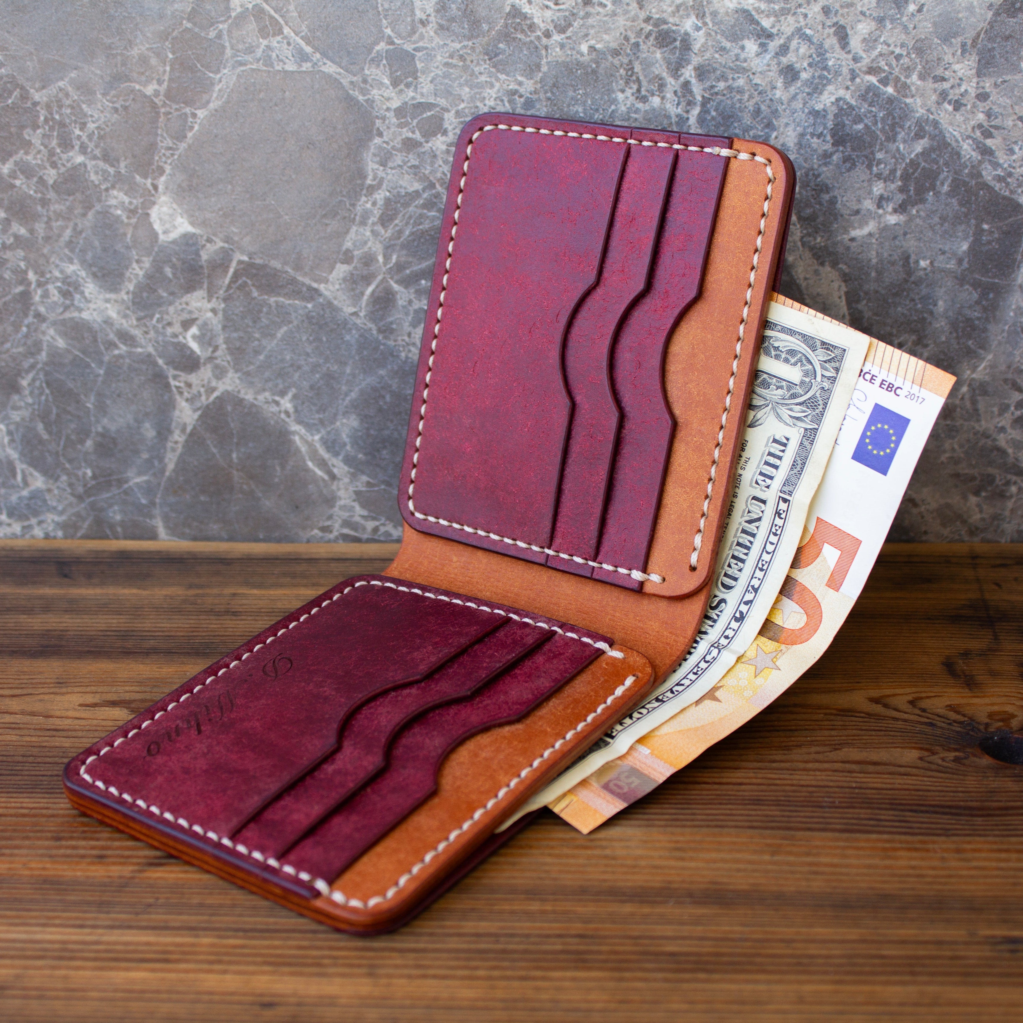 A two-tone leather card holder wallet in burgundy and tan, shown open with Euro banknotes tucked into the card slots, set on a wooden surface against a gray marble background.
