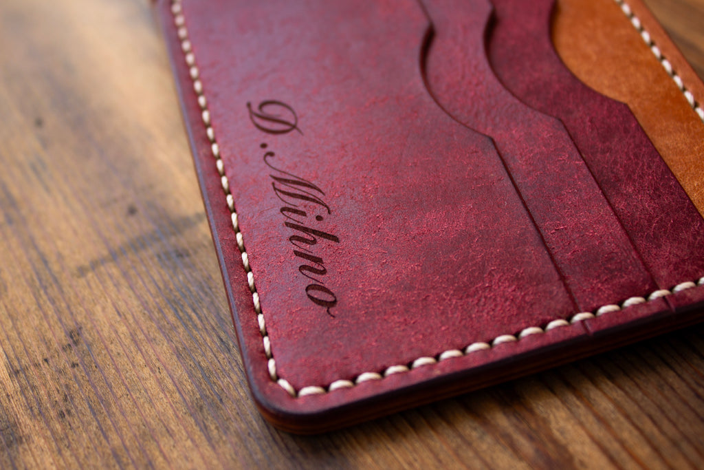 A close-up of a dark red leather cardholder wallet with white stitching and an engraved cursive logo, resting on a wooden surface.