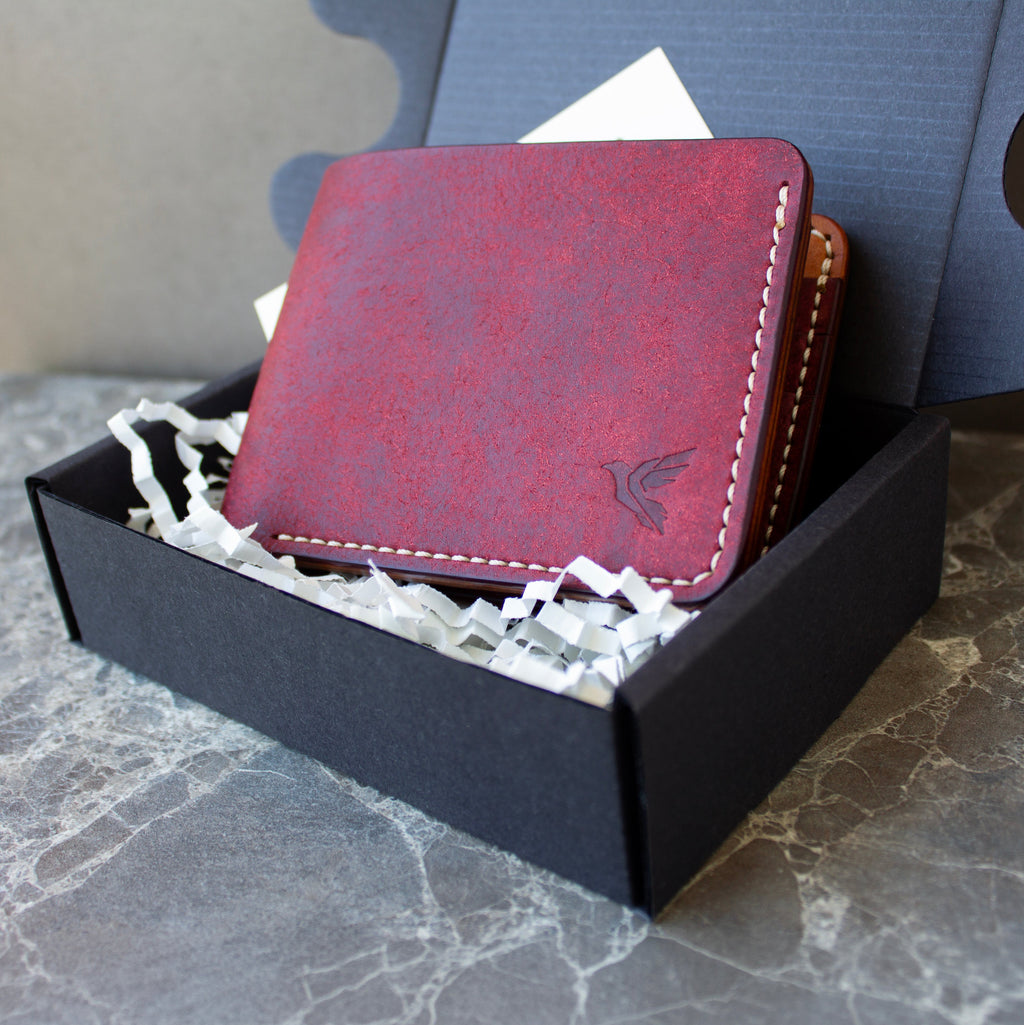 A red leather bifold wallet with white stitching and an embossed bird logo, presented inside a black gift box with white paper shred on a gray marble surface.