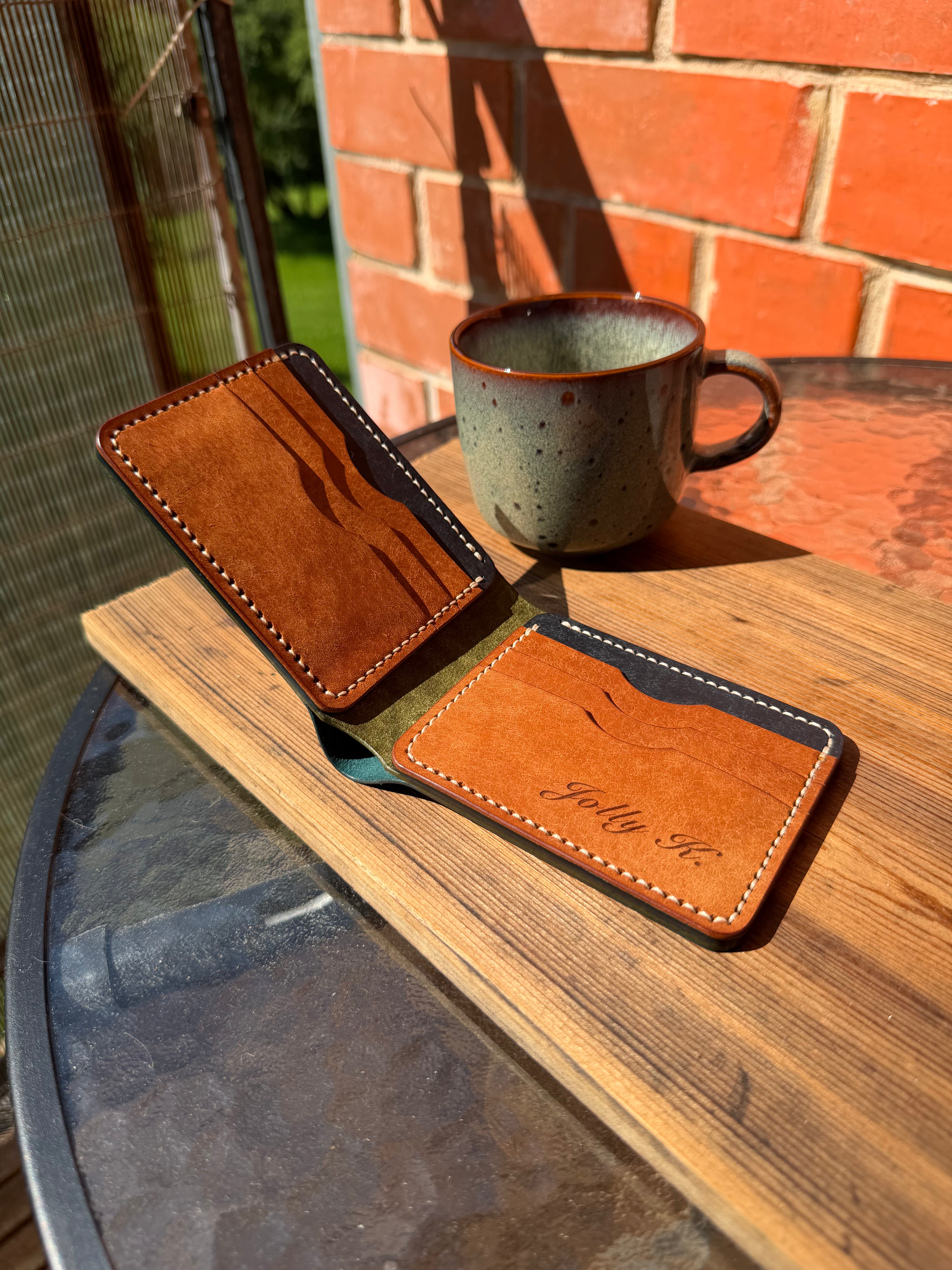 A brown leather card holder wallet with white stitching and an engraved name, displayed on a wooden table next to a ceramic mug in an outdoor setting.