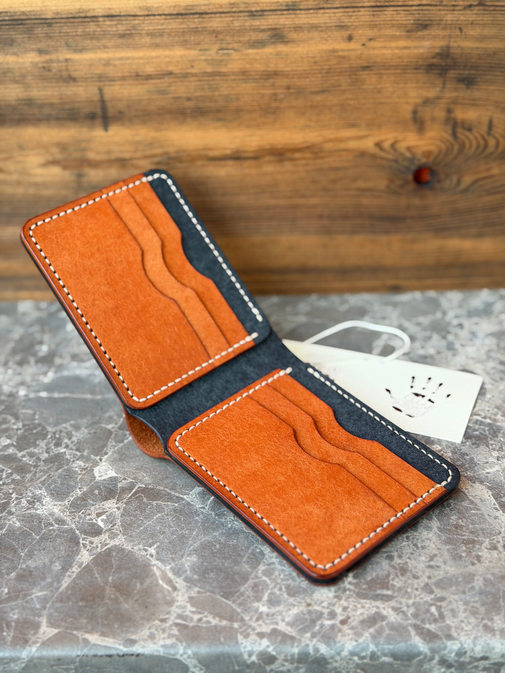 An open minimalist bifold wallet made of textured orange and dark gray leather, displayed on a gray marble surface with a wooden background and a small white tag.