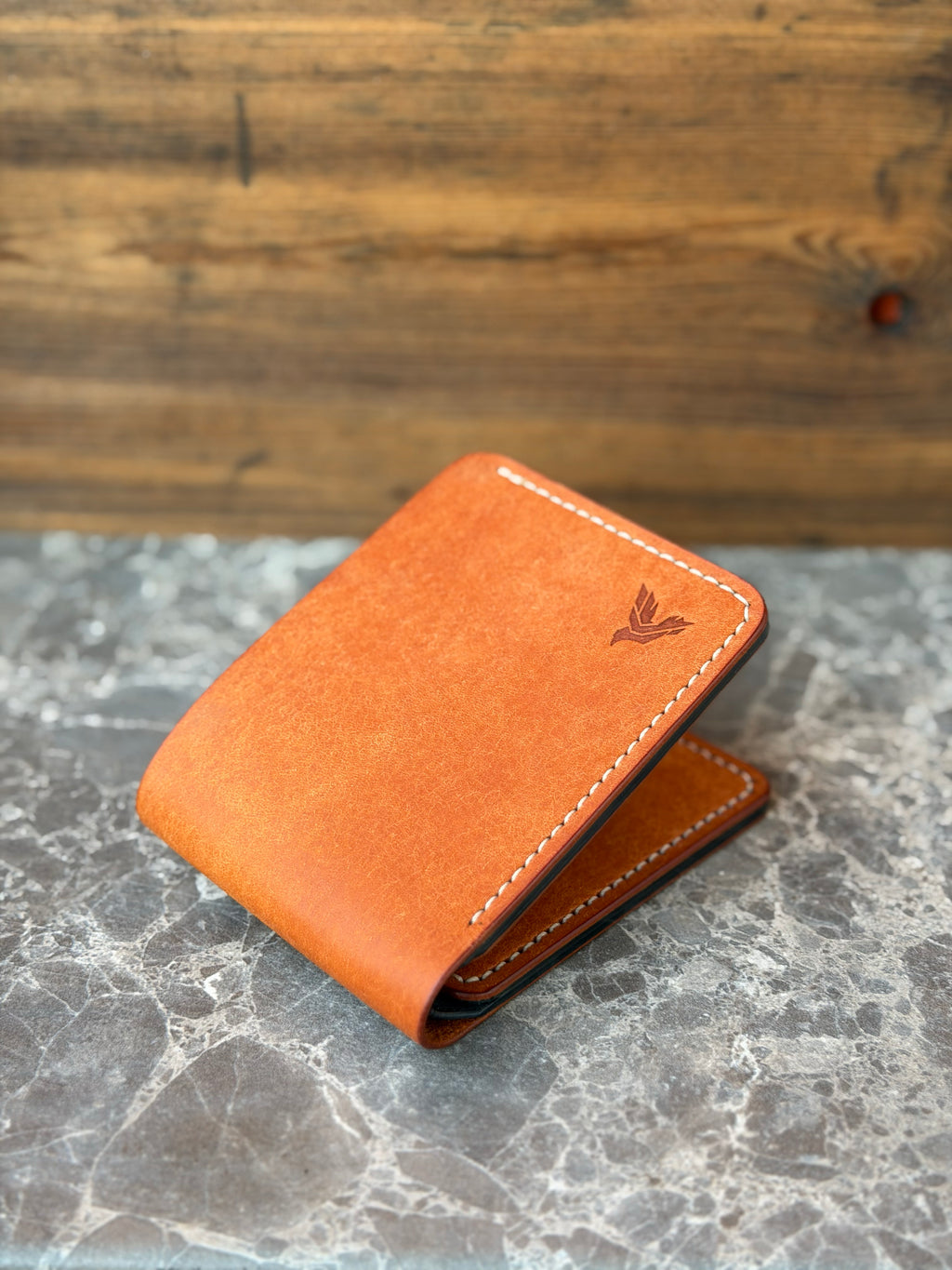 A slim, tan leather bi-fold wallet with white contrast stitching and an embossed bird logo, resting on a gray marble surface with a rustic wooden background.