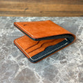 A slim brown leather bifold wallet with white contrast stitching, shown open to reveal a dark navy interior and card slots, resting on a gray marble surface with a wooden background.