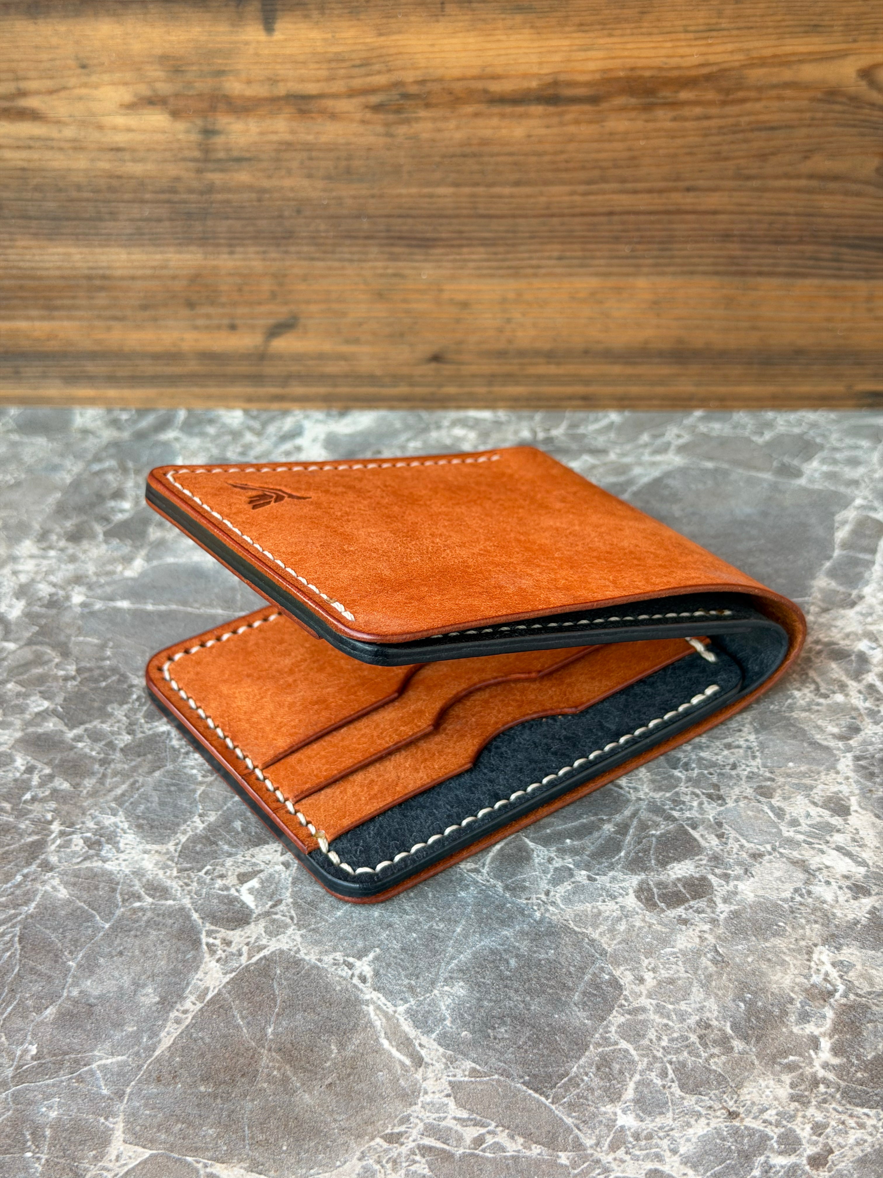 A slim brown leather bifold wallet with white contrast stitching, shown open to reveal a dark navy interior and card slots, resting on a gray marble surface with a wooden background.