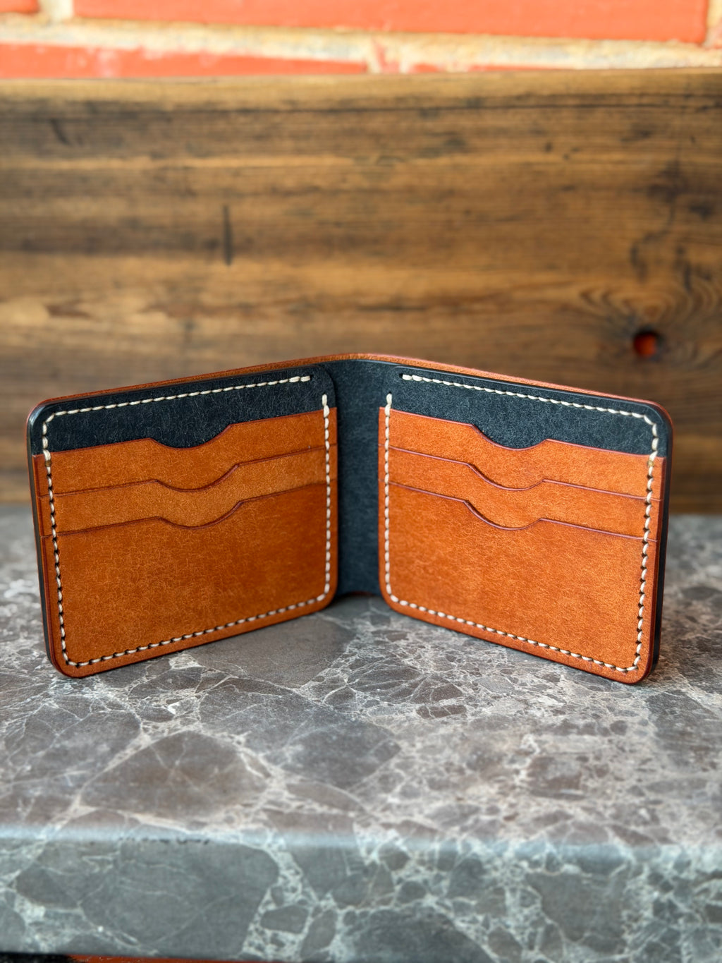 A brown leather bifold wallet with card slots and contrast stitching, shown open on a gray marble surface with a rustic wooden background.