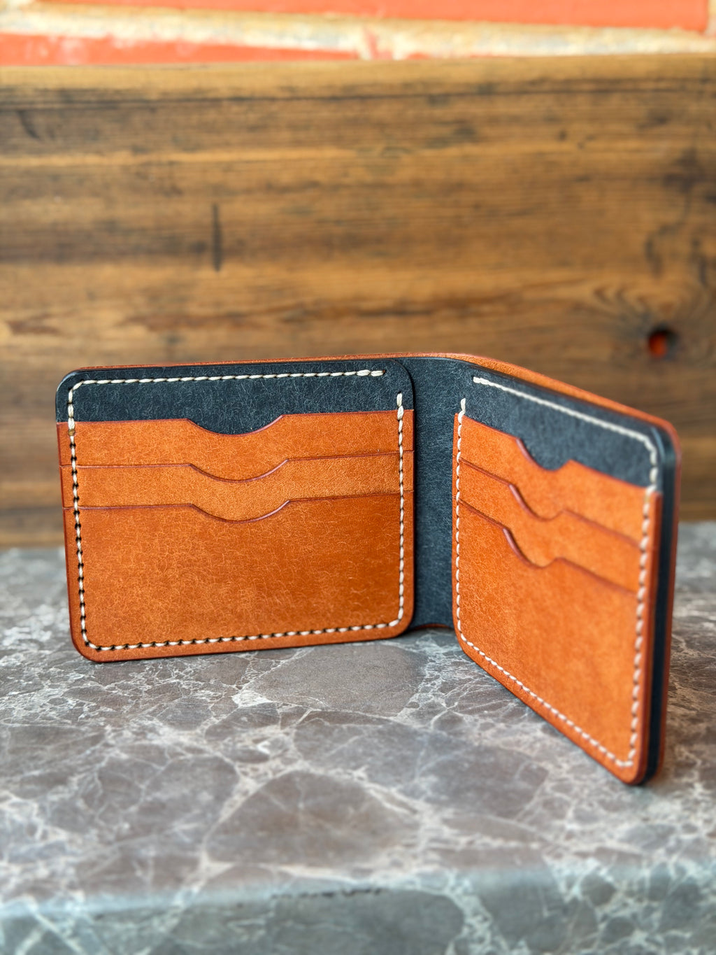 An open bifold leather wallet in warm tan and dark charcoal gray, displayed on a gray marble surface with a rustic wooden background.