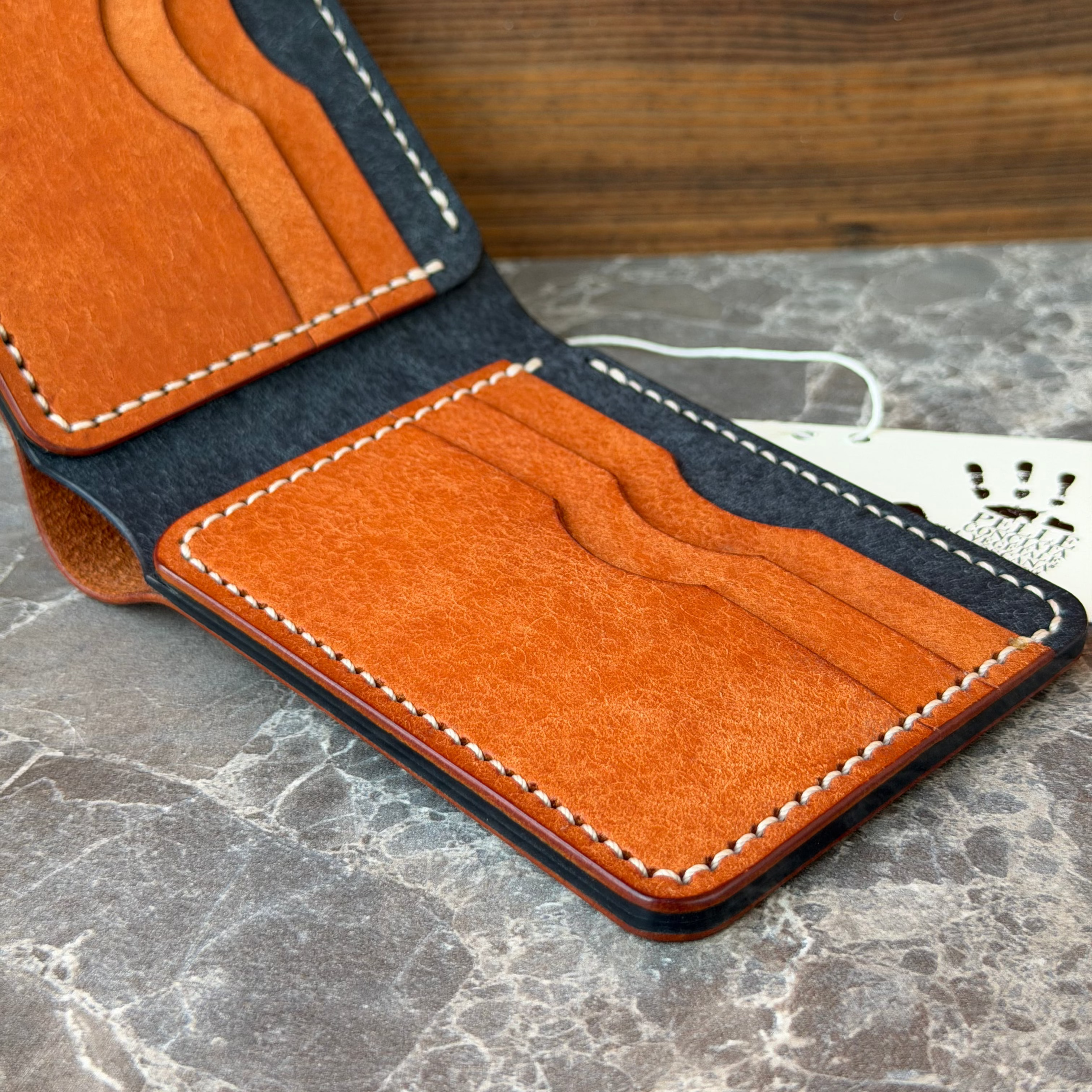 An open minimalist leather cardholder wallet in orange suede-like material with dark navy blue lining and white contrast stitching, resting on a gray marble surface with a wooden background.