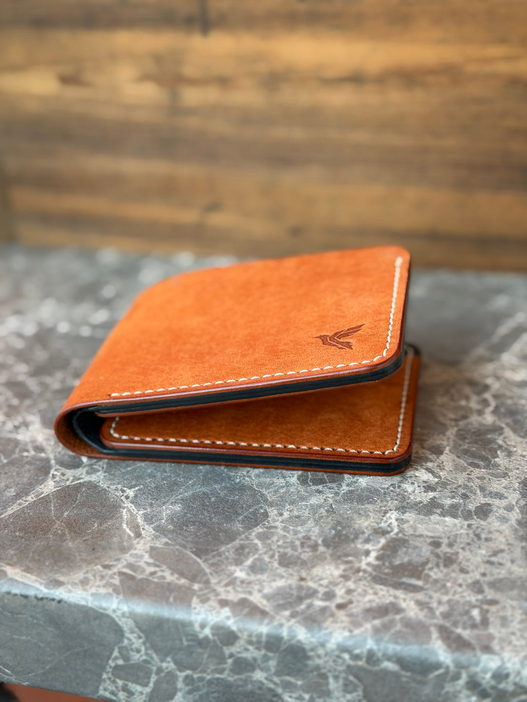 A slim, orange-brown leather wallet with white contrast stitching and a debossed bird logo, resting on a gray marble surface with a blurred wooden background.