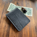 Black leather wallet on a wooden surface with a dollar bill and a pen.