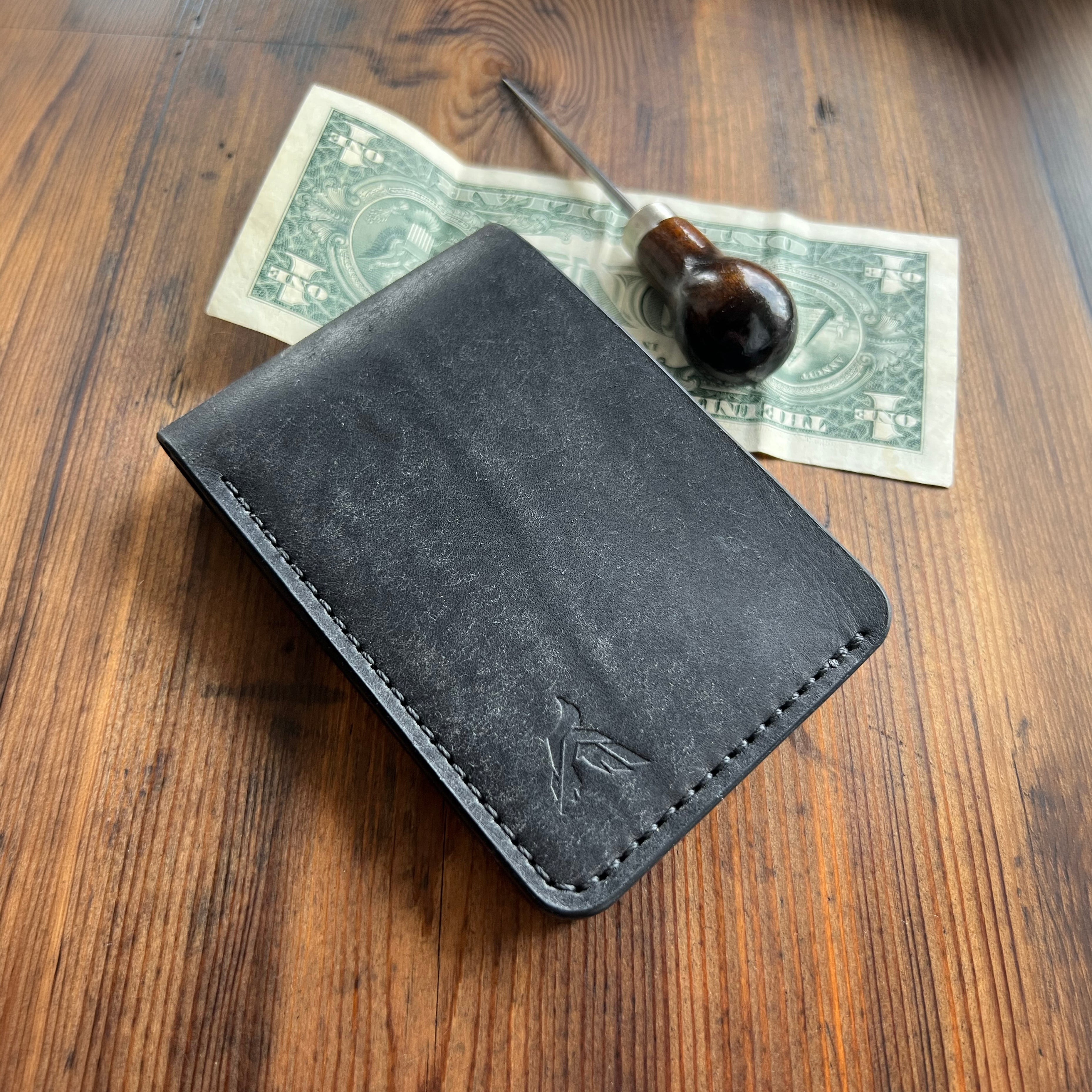 Black leather wallet on a wooden surface with a dollar bill and a pen.