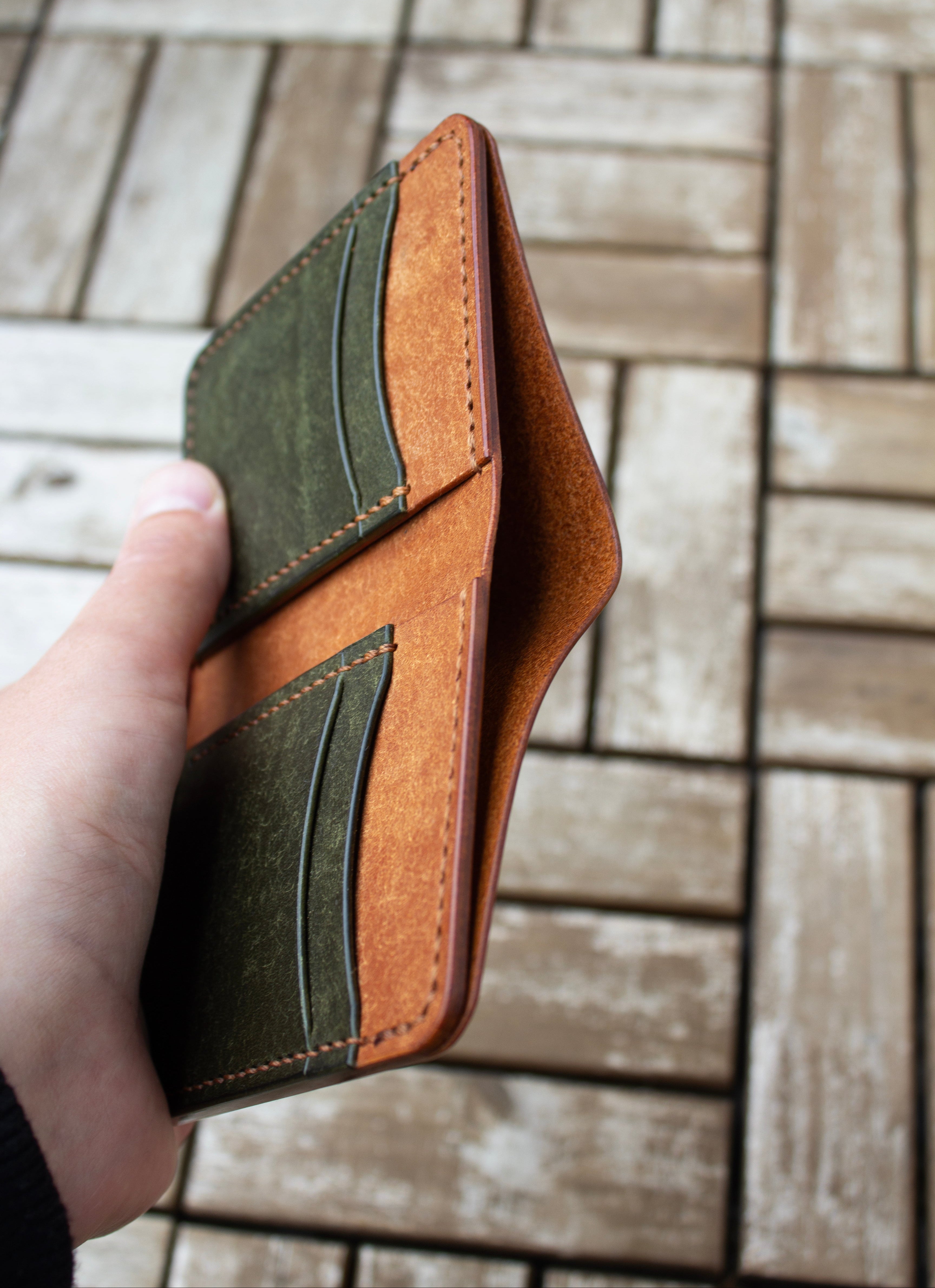 Hand holding a brown leather wallet with green accents on a tiled floor.