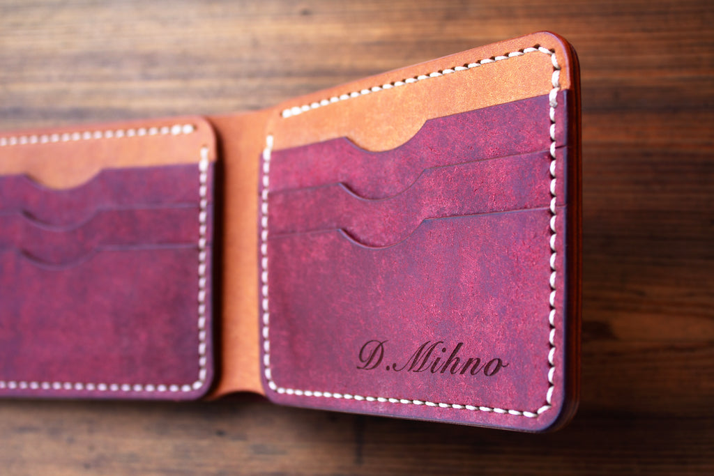 A close-up of an open leather bifold wallet on a wooden surface, showing a burgundy textured interior with card slots, white stitching, and the name 'D. Mihno' embossed on the right panel.