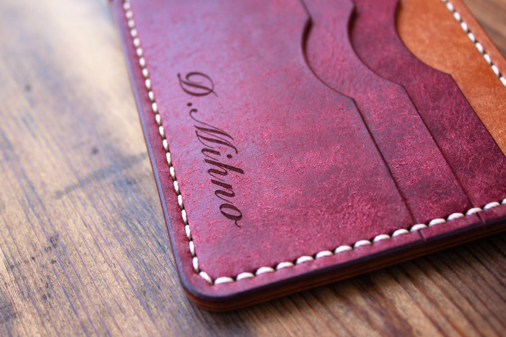 A close-up of a reddish-brown leather cardholder wallet with white stitching and an engraved cursive name, shown on a wooden surface.