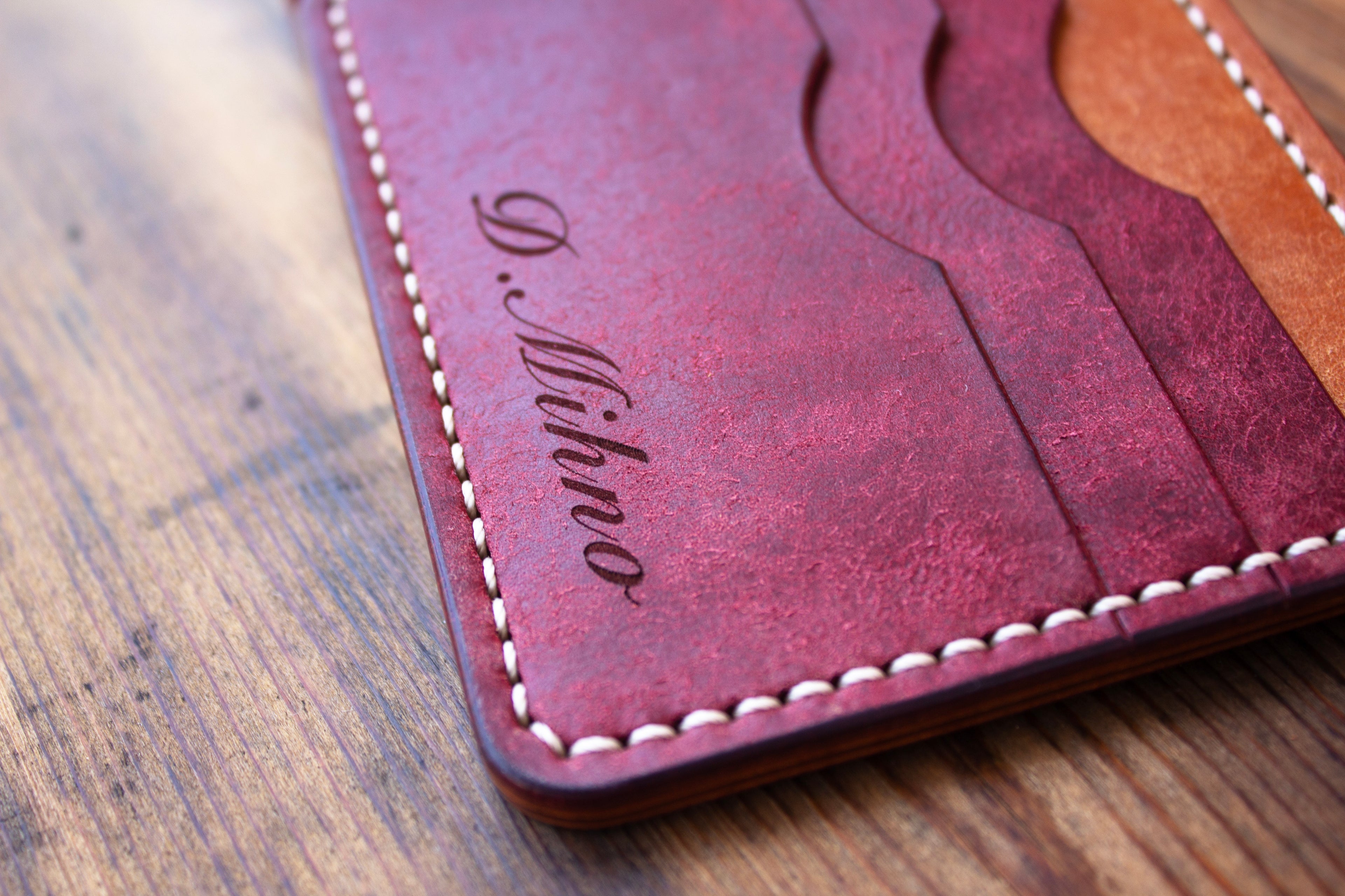 A close-up of a reddish-brown leather cardholder wallet with white stitching and an engraved cursive name, shown on a wooden surface.
