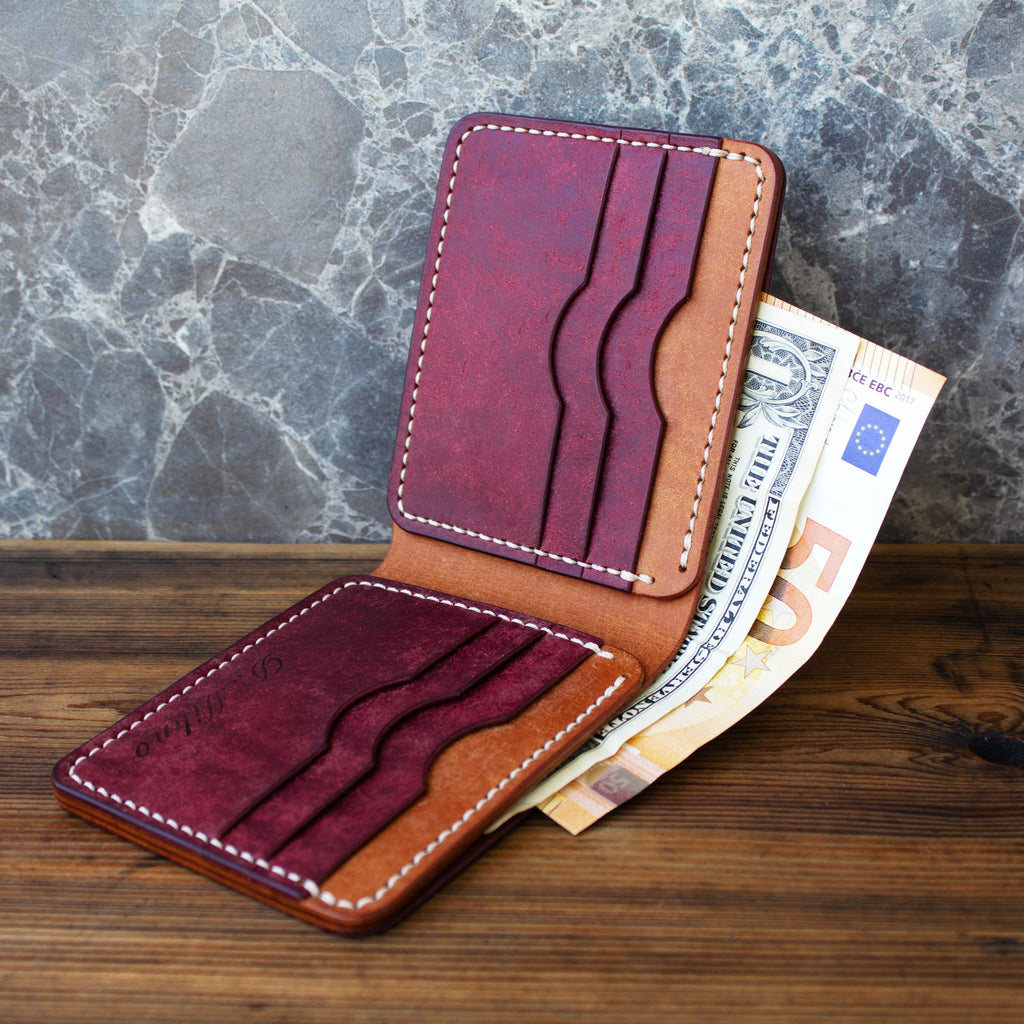 A slim brown leather card holder wallet shown open on a wooden tabletop, with burgundy card slots and white contrast stitching. Euro banknotes are tucked inside, and the wallet is photographed against a gray marbled wall.