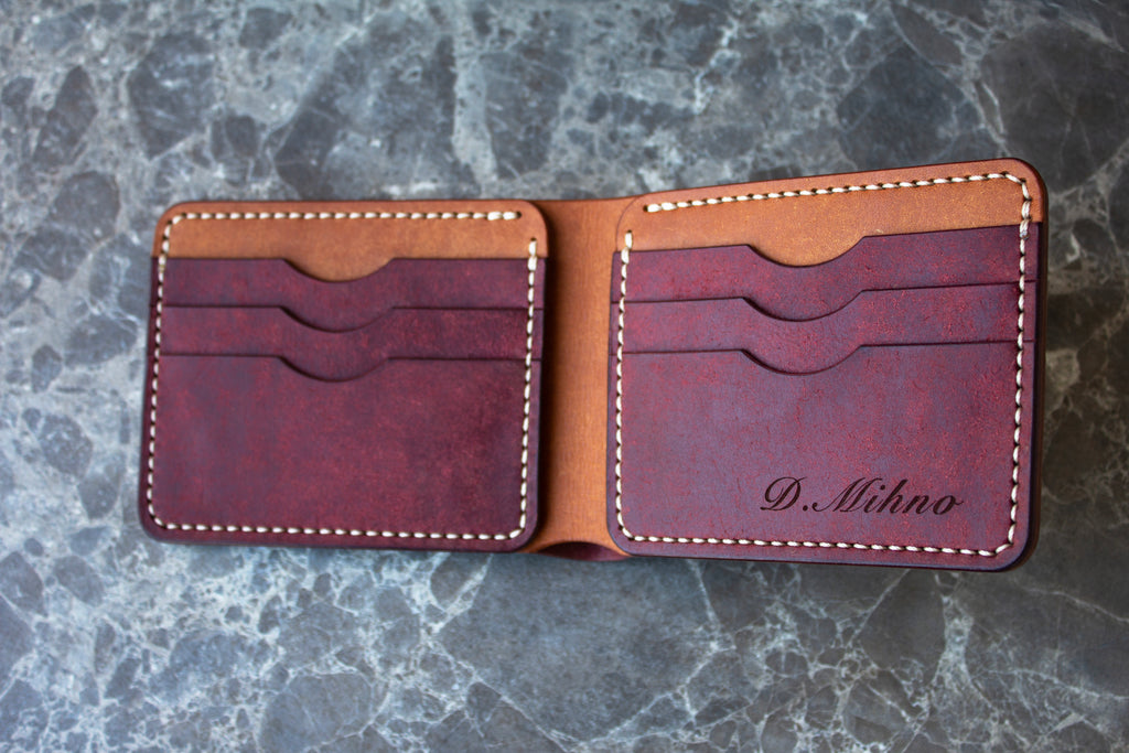 An open bifold leather wallet in a deep burgundy and tan color combination, featuring white contrast stitching and the name 'D. Sihno' embossed on the right pocket, displayed on a gray marble surface.