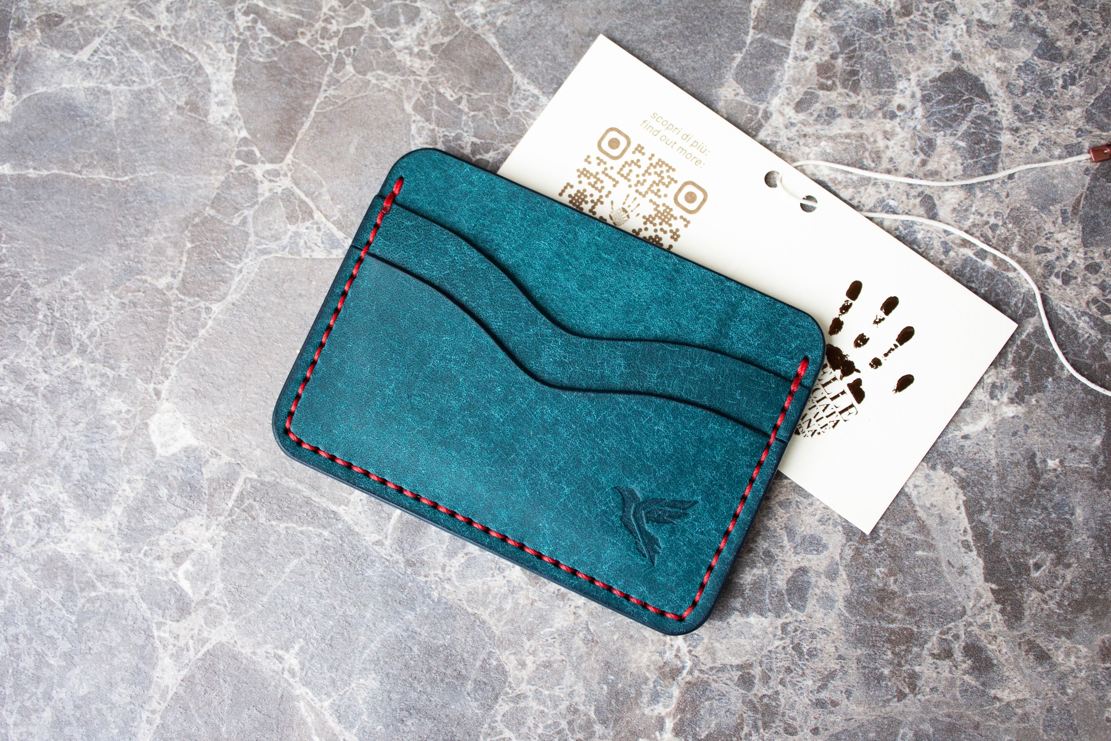 Teal cardholder with red stitching on a gray surface with a white card and brown design.