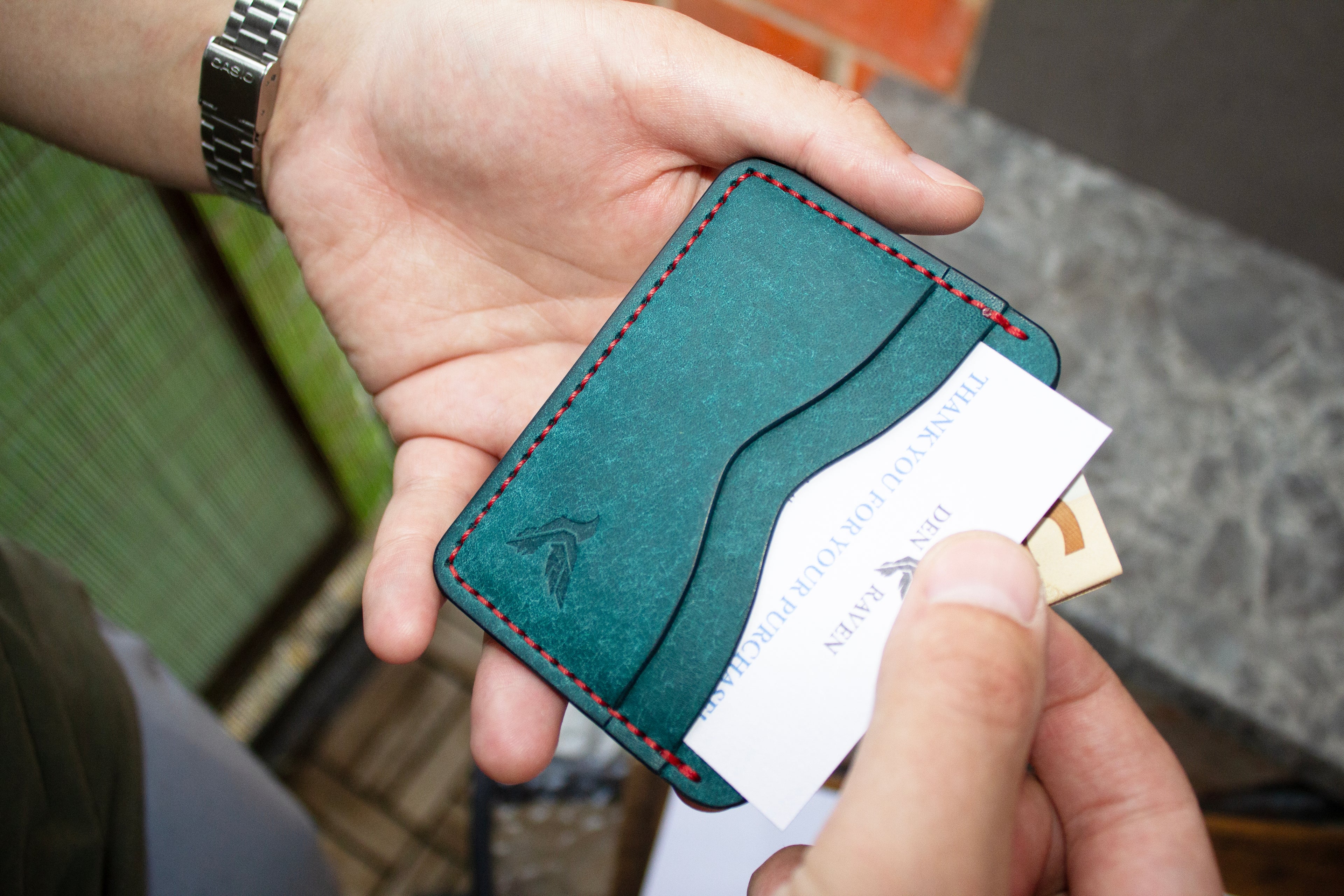 Hand holding a teal leather cardholder with red stitching
