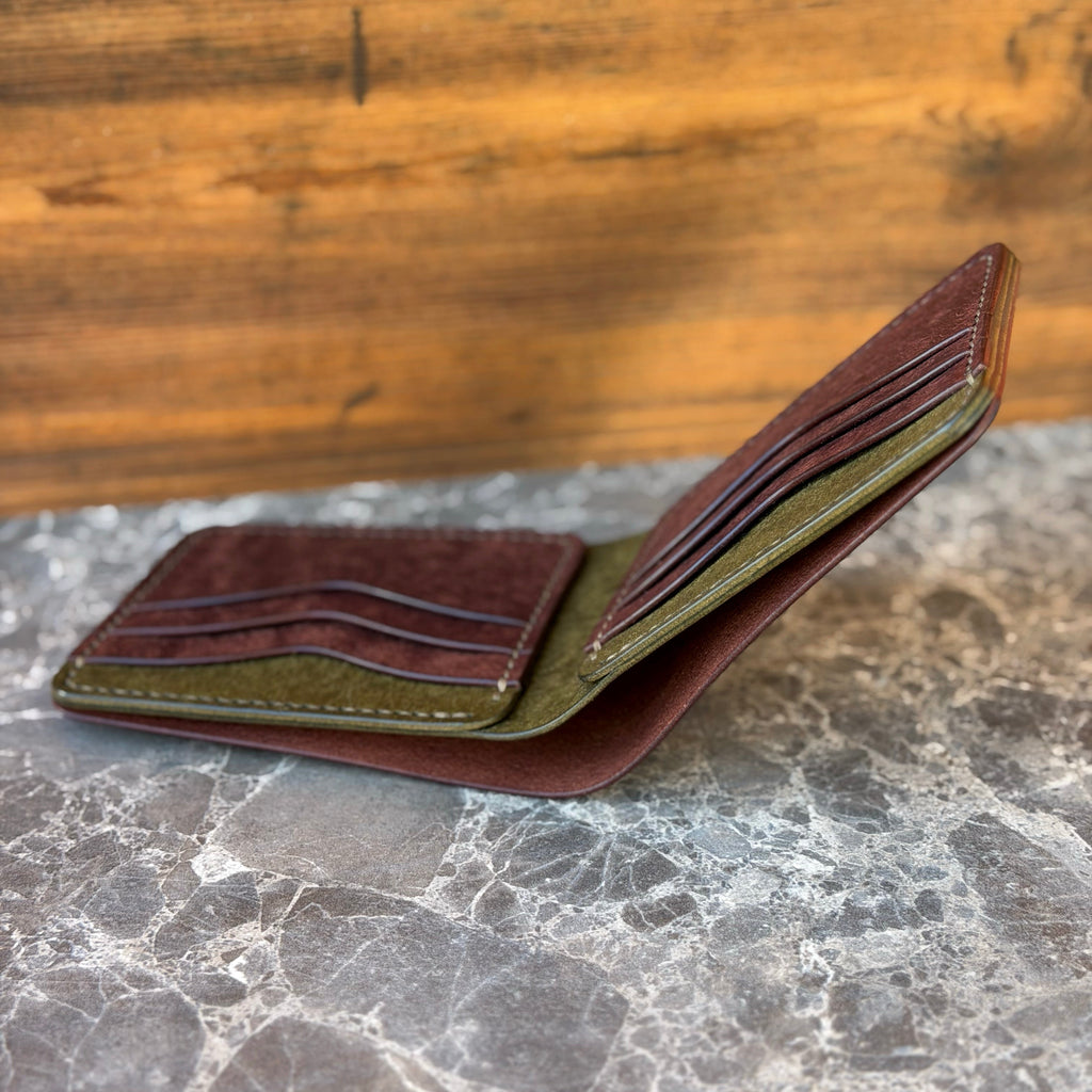 A brown leather bifold wallet shown open, displaying its interior card slots, dark brown card protectors, and an olive green inner lining, resting on a gray marble surface with a wooden background.