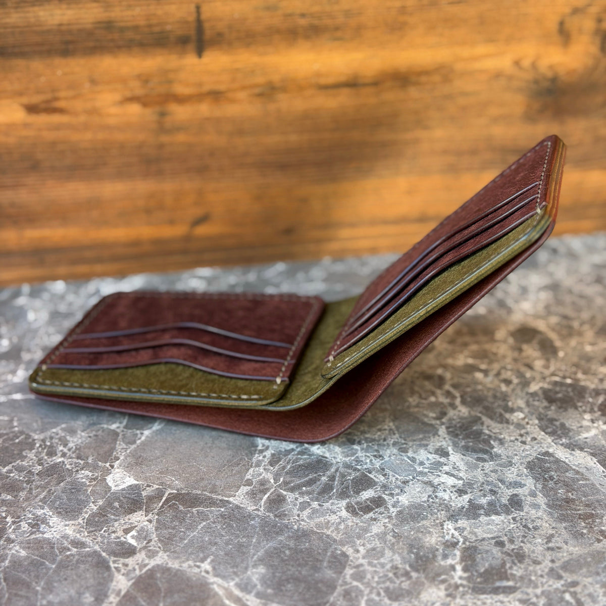 A brown leather bifold wallet shown open, displaying its interior card slots, dark brown card protectors, and an olive green inner lining, resting on a gray marble surface with a wooden background.