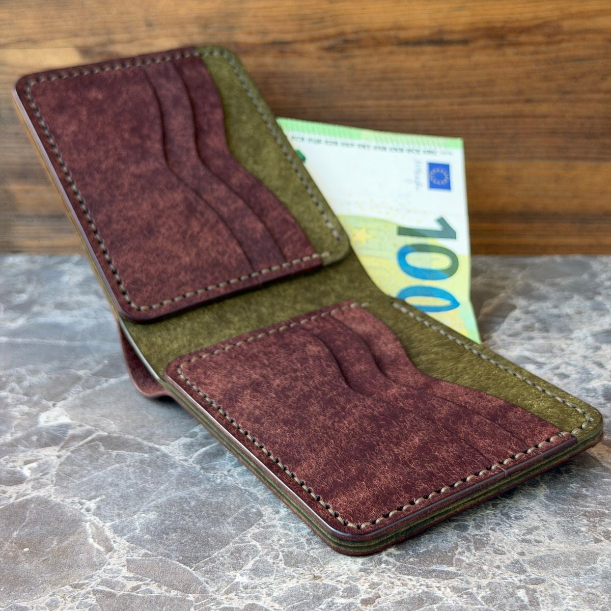 A minimalist bi-fold wallet made of burgundy suede and olive green leather, shown open with a 100 Euro banknote tucked inside, set on a gray marble surface against a wooden background.