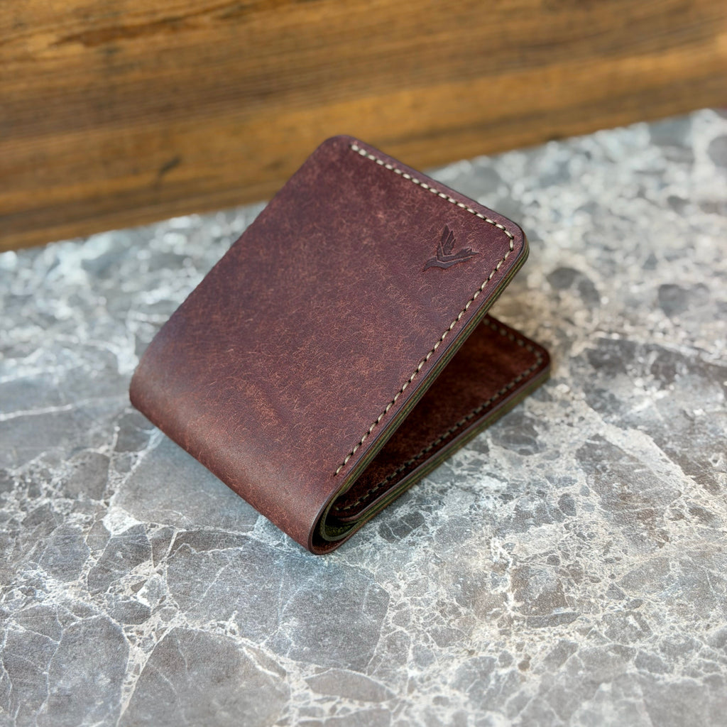 A dark brown leather bifold wallet with visible stitching and a small debossed logo, resting on a gray marble surface with a wooden background.