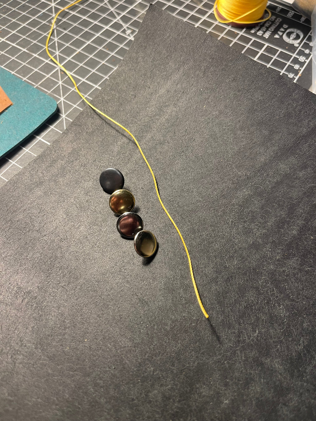 A close-up of a dark gray felt surface with four small round cabochon embellishments in black, bronze, brown, and gold tones, alongside a yellow thread and a gridded cutting mat in the background.
