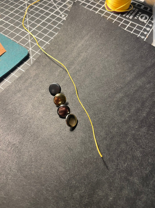 A close-up of a dark gray felt surface with four small round cabochon embellishments in black, bronze, brown, and gold tones, alongside a yellow thread and a gridded cutting mat in the background.