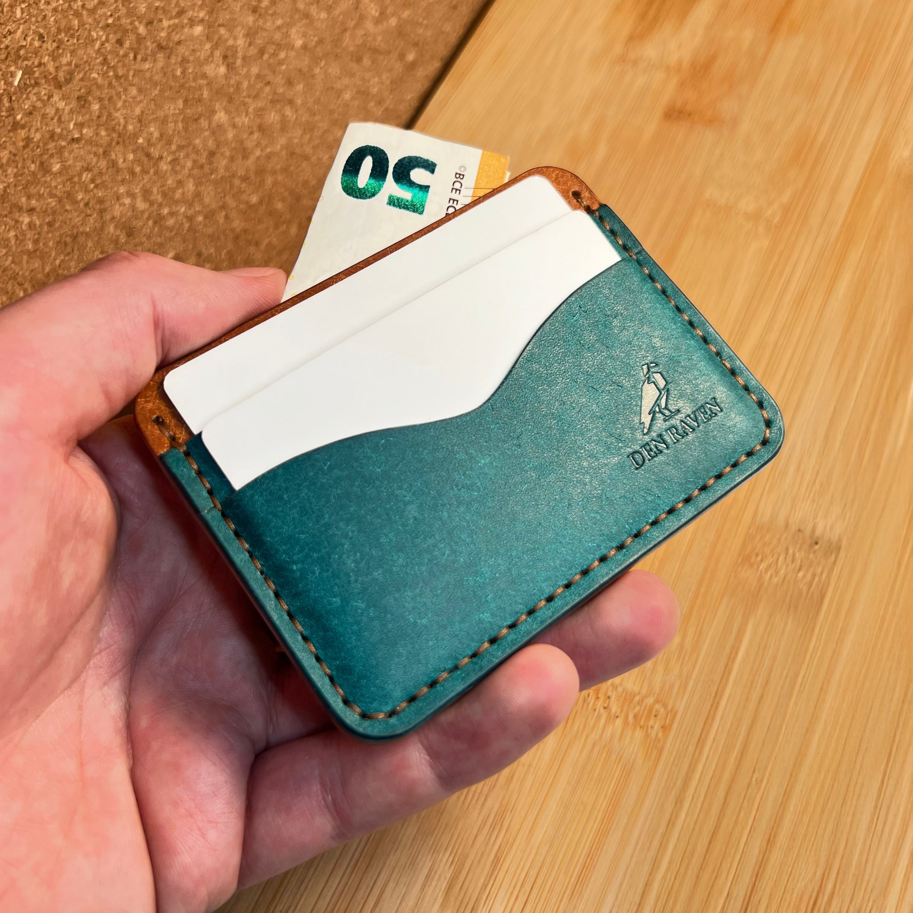 Hand holding a teal leather wallet with a card and money on a wooden surface