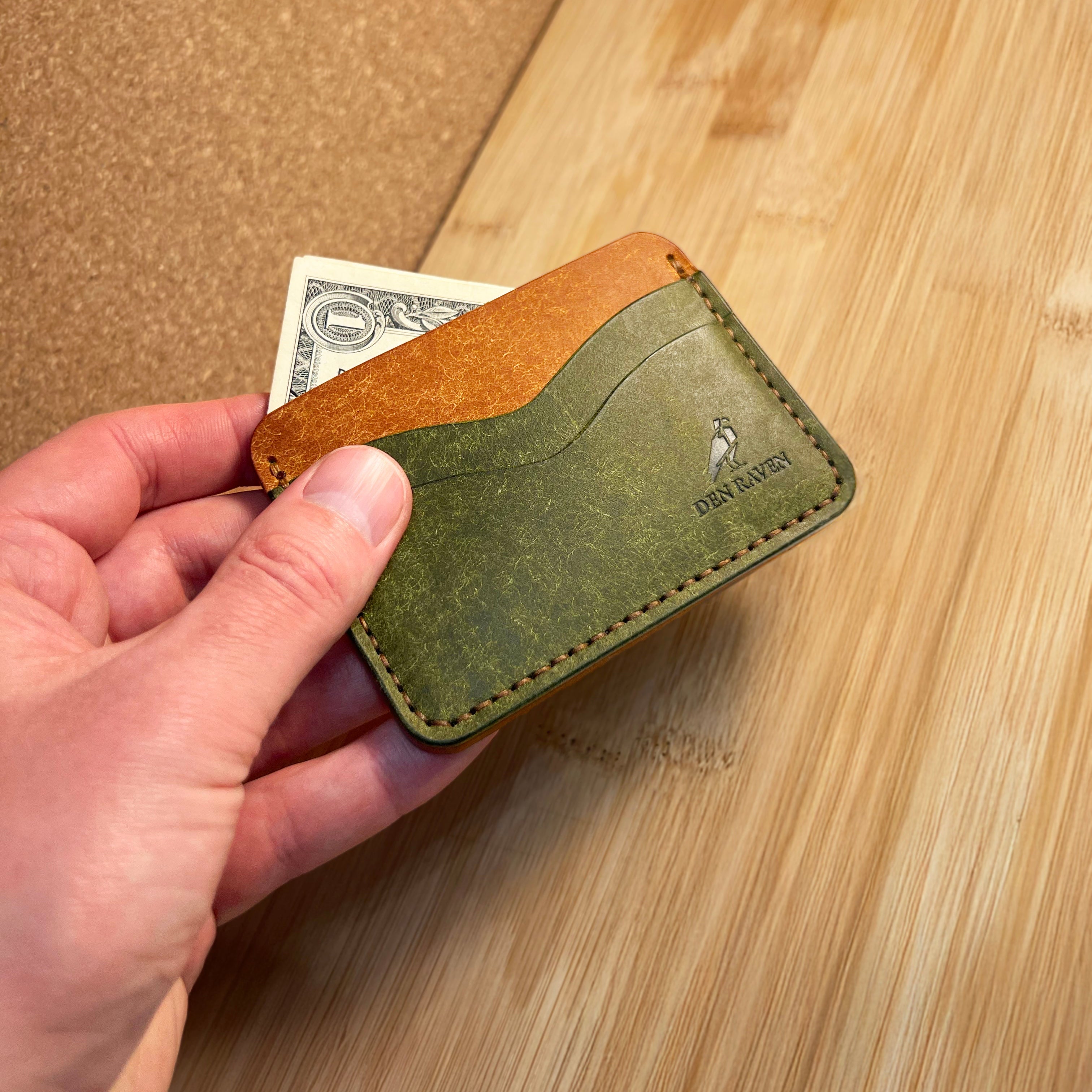 Hand holding a green leather wallet with a brown flap on a wooden surface