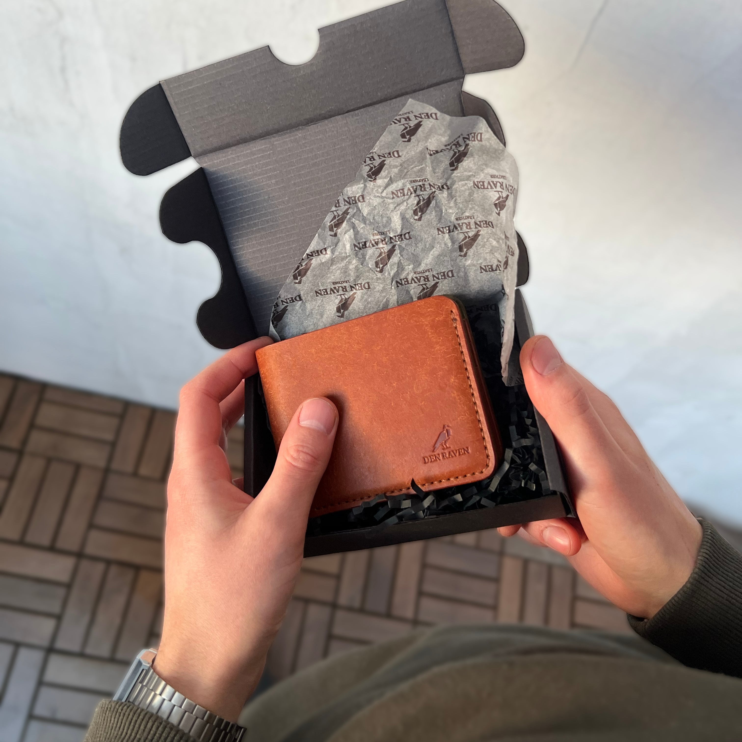 Person holding a brown leather wallet with a black box on a light gray background