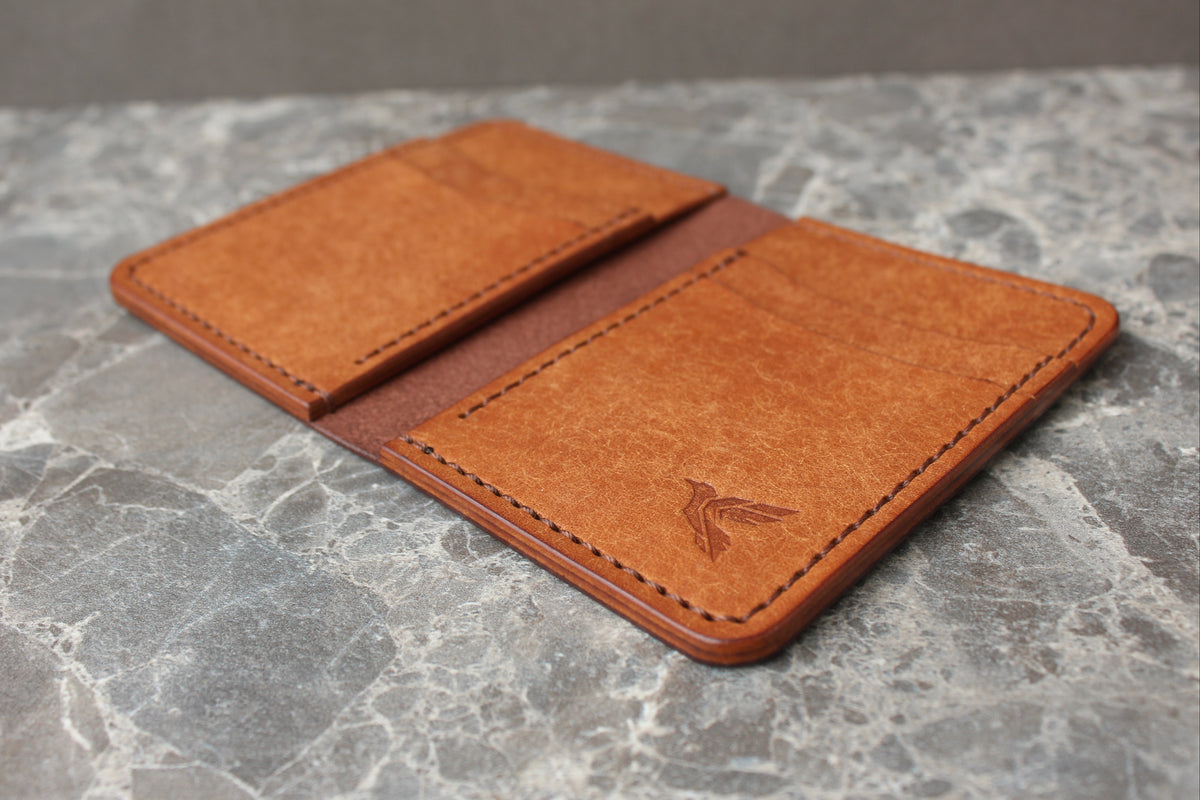 A tan leather card holder wallet with a stitched border and an embossed bird logo, displayed open on a gray marble surface.