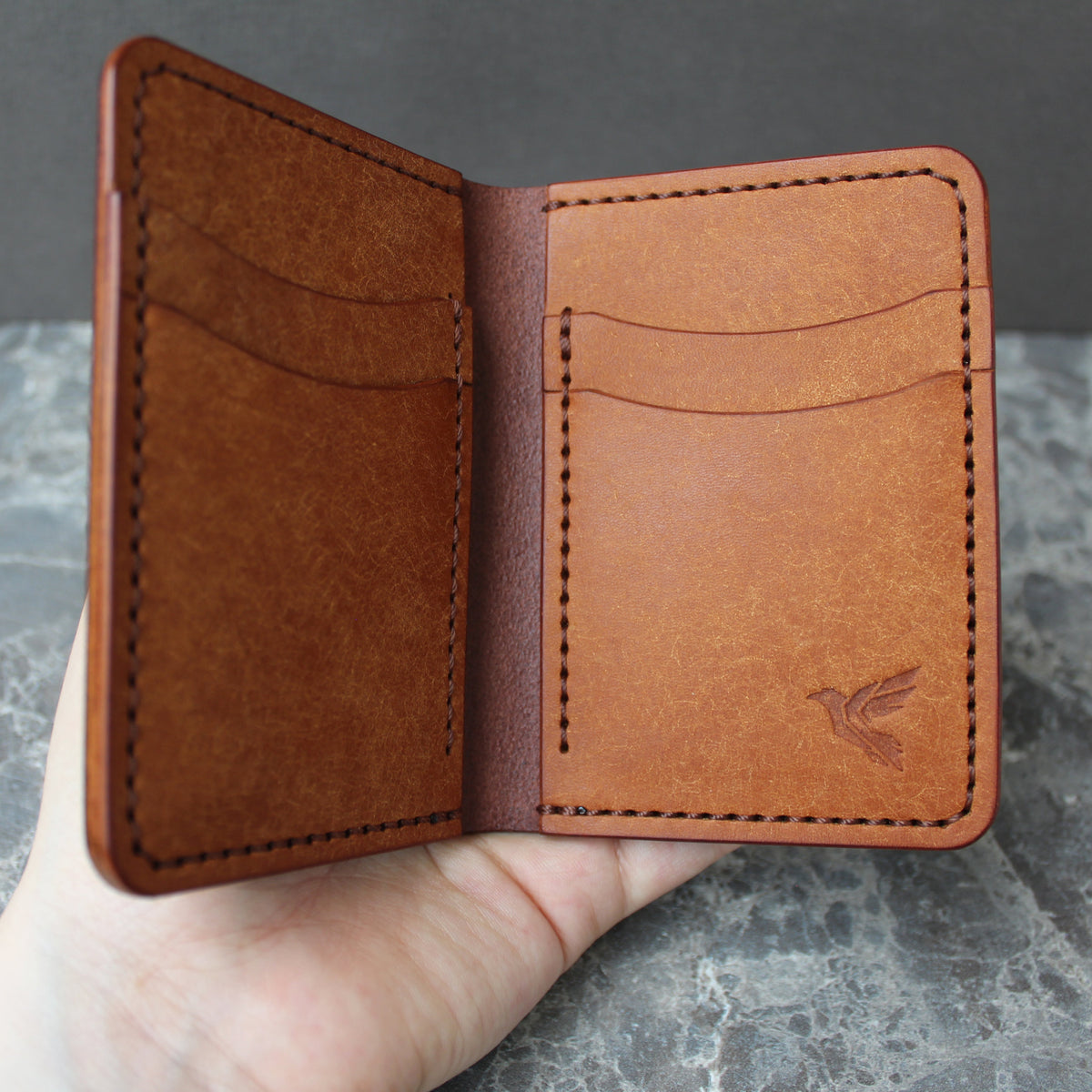 A hand holding an open brown leather bifold wallet with card slots and a debossed bird logo on the right panel, set against a gray marble surface and dark gray background.